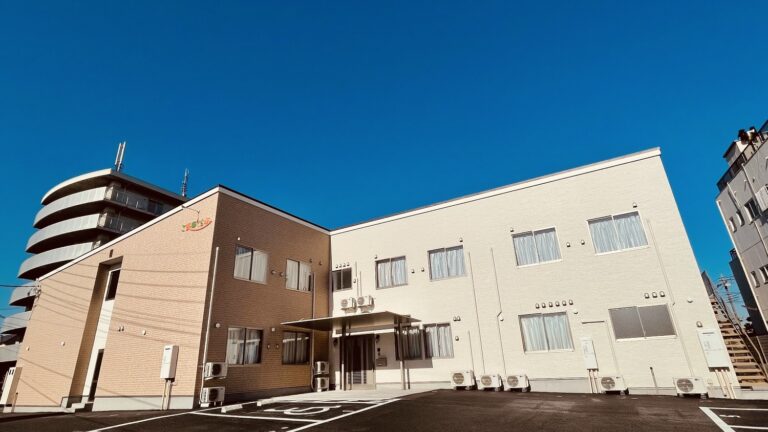介護施設の建物外観。明るい色の外壁と駐車場が見える。